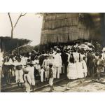 Four drillers stand under a thatched rig while a group of people look on.