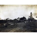 A photo of a boy walking down a road towards an oil derrick covered in thatching. There is a large crowd underneath the derrick. The area beside the road is empty and there are trees in the background.