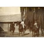 A photo of two boys on horses with a man in the middle riding a horse. There is a building covered in metal siding and roofing behind them, as well as a tree with long, hanging branches.