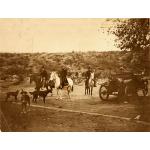 Trois hommes à cheval. Il y a trois chiens devant eux et un long tuyau qui est à moitié enfoui. Un quatrième homme est assis dans une voiture à droite. On aperçoit une colline avec des buissons à l'arrière-plan. 