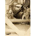 Four men stand inside of an oil rig that is covered with sheet metal. There is a large pipe running horizontally a foot from the floor.