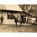 A photo of a man wearing a suit riding a dark horse. He is holding a whip in his hand. There is a building with siding and a covered porch behind him. Another man leans against the wall of the building.