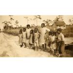 A photo of a group of children standing beside a dirt road. There is a building behind them and trees in the background.