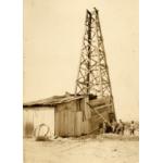 Four men standing in a dusty field beside a wooden oil rig with four sides. The bottom of the rig, with the machinery inside it, is covered in wooden boards and metal sheeting.