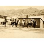 A photo of nine International Drillers sitting on horses and wearing hats. There are two single-story buildings behind them and rolling hills in the background.