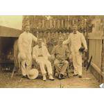 Two men sitting in chairs with a man standing on either side. They are wearing white and light coloured clothing. Each man is holding a hat or resting one on the ground. They are standing in front of a fence with vines on it. 