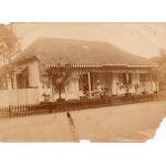 Three people standing outside of a house. There are trees to the sides and plants on the veranda. There is a dark picket fence at the top of a steep wall that leads down to a moat. 