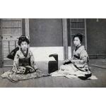 A photo of two girls dressed in Japanese clothing, sitting on folded mats. There is a dark box sitting open between them. There is a wall with matting behind them. 