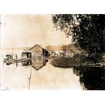 A photo of three small structures sided with reeds on a raft close to the shore of a river. There are people standing on the raft. There is a small boat to the left and reeds and a tree to the right. 