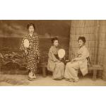 A photo of three girls holding fans, dressed in Japanese clothing. One is standing and the other two are sitting on a wooden bench. There is a reed mat against the wall and an illustrated scene of waves behind them. 