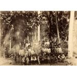 Photo d'un groupe d'écoliers avec quatre adultes au dernier rang. Ils sont sous un arbre avec de grandes feuilles et il y a des arbres derrière eux.  