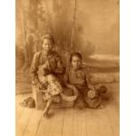 A photo of an old woman sitting on a wooden box with her legs crossed. A younger woman sits on the floor made of wooden boards beside her. There is a screen behind them with trees on it.  