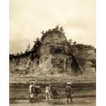 A photo of four people with two rickshaws standing on a road in front of a tall cliff with trees on the top of it. 