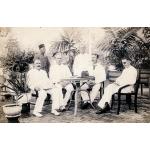 A photo of five International Drillers, wearing white, around a table with a hat on it. A servant stands behind them. There are trees in the background. 