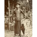 A photo of a Chinese man wearing a hat, buttoned shirt, and skirt. He is holding a closed umbrella at his side. There is a structure with wooden poles and a roof behind him.