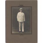 William McRae debout vêtu d’un costume blanc et d’un chapeau Panama. La photographie est placée dans un papier marron mat. 