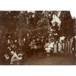 William McRae sitting in a white suit and pith helmet, surrounded by a group of locals under an awning. 