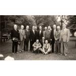 A group of retired International Drillers. Eleven are standing and three are sitting in front on grass. They are all wearing suits. There are trees behind them.