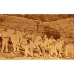 A photo of two rows of International Drillers, all wearing white, sitting in front of a long log that is taller than the men.