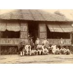 A group of people standing and sitting in front of a thatched bungalow. The doorway is open and there is a thatched window propped open on either side of the doorway.