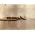 A photo of a group of men pulling a rope attached to a boiler down a stone pier that juts out into the water. There are four poles set up like a pyramid at the end of the pier.