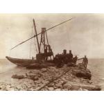 A photo of a boat in the water at the end of a stone pier. There is a crane lifting a steam boiler onto the rocks, and a man walks on the edge of the pier watching the boiler.