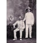 Une photo de deux hommes devant une toile de fond sur laquelle des arbres sont peints. Ils portent des costumes blancs. Une table se trouve à côté de l'homme assis à gauche, et des livres et des fleurs y sont disposés. L’homme à la droite porte un chapeau blanc.