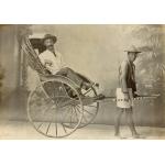 Une photo d'un homme assis dans un pousse-pousse à deux roues avec l'auvent refermé. Il est tiré par un homme de la région qui porte un chapeau de paille. Une toile de fond peinte se trouve derrière eux, avec des piliers et des arbres. 