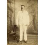 A photo of Thomas Knapp in front of a painted background of trees. He is wearing white and is resting his hand on the back of a chair with a hat on the seat.