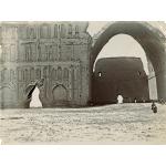 A photo of a group of people in front of a crumbling stone building. There is a giant archway on the left that rises three stories in the air. The wall on the left is carved with arches and pillars and there is a large triangular hole at the base.