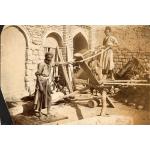 A photo of four men standing in front of a stone wall with arches in it. Two of them using a saw to cut a piece of wood that has been propped up in a wooden frame.