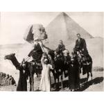 A photo of Mr. and Mrs. Saunders and their son riding camels. There are three local people standing on the ground and holding bottles on their heads. The Sphinx and a pyramid are in the background. 