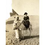 Une photo d'Annie Dyke dans le désert, assise sur un âne blanc tenu par un Égyptien. Elle porte une robe à rayures foncées et un chapeau, tandis qu'il est vêtu d'une robe blanche. Il y a une pyramide en arrière-plan. 