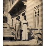 A photo of two men wearing long robes with dark hats and bands around their waists. They are standing beside a bench in front of a striped stone building.