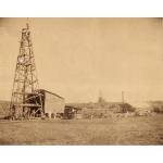 A photo of several International Drillers, wearing white suits and pith helmets, stand in front of an oil derrick in a grassy field. There is a small building and another oil derrick in the background.