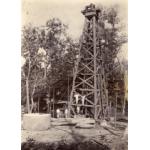 A photo of a group of International Drillers standing at the base and on the first support beam of an oil derrick. They are wearing white. There is a large cylinder and some logs in front of them and trees in the background.