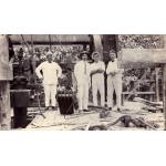 A photo of four men standing inside an oil derrick on a wooden plank floor. They are wearing white. There is a large wheel behind them and machinery beside them.