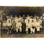 Group photograph of foreign drillers in Borneo all wearing white suits. They are sitting or kneeling on a raised platform underneath a roofed structure. 