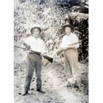 Two men in white shirts, hats, and dark pants. They are holding rifles. There is a small dog at the foot of the man on the right. They are standing in front of a wall of vegetation.