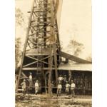 A group of men standing in front of a wooden oil rig. There is a ladder on the front side of the rig. The machinery beside the rig has a metal sheet roof over it.