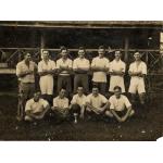 A photo of a group of men wearing white shirts. Seven are standing with their arms crossed. Five are sitting cross-legged with a ball on a stand in front of them.