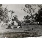 Four people in front of a canopy tent. Three are sitting on logs and the man on the right is standing. The woman beside him is wearing a white skirt. There are trees in the background.