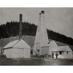 A photo of three men standing in front of an oil rig covered in wooden planks. There is a white building with a door at the front and a chimney coming out of the roof on the left. There is a line of trees in the background.