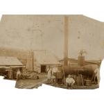 A photo of four men standing in front of a steam boiler. There are two other people off to the side. There is an oil rig in the background. The bottom of the rig is covered in wooden planking.