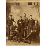 A photo of six men in front of a painted backdrop. They are wearing dark suits and two are sitting in chairs. There is a patterned rug underneath their feet.