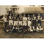 Twelve men wearing soccer uniforms in front of a net. Eight of the men are standing (seven with their arms folded) and four are sitting. There is a ball in the middle of the seated players.  