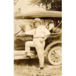 Une photo de Fred Webb debout, le pied sur le marchepied d'une voiture. Webb porte une chemise et un pantalon blancs, une cravate et un chapeau de plaisancier. La voiture a un toit en toile et aucune fenêtre. Il y a un bâtiment avec un toit en pointe à l'arrière-plan. 