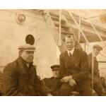 Photo de quatre hommes assis au pied d'un escalier métallique sur le pont d'un bateau. Ils portent des vestons foncés. 