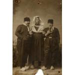 Une photo de trois hommes chaussés de sabots. L'homme du milieu porte une jupe ainsi qu'un foulard sur la tête. Les hommes de chaque côté portent des pantalons à taille haute avec deux boutons et des foulards noués autour du cou. Ils ont tous les deux une pipe.  