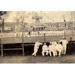 A photo of seven men sitting on the outside of a curved wooden track. There is a large group of men inside the track, and one has a bicycle. 